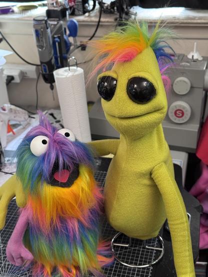 A green alien hand and rob puppet with large glossy black eyes and a rainbow Mohawk with its arm around a small rainbow fur hand puppet