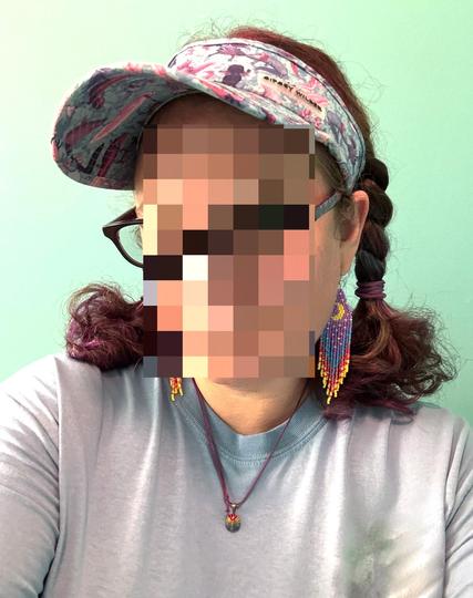 A selfie of a person with their face blurred wearing a light blue crew neck top, a sun visor, pigtail braids with streaks of color, a pendant necklace, and a pair of beaded fringe earrings with crescent moon shapes on them