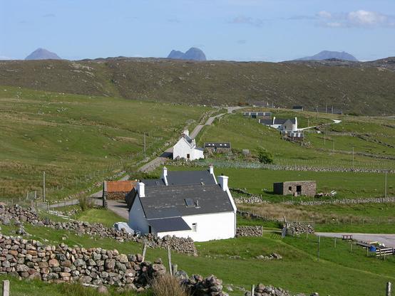Clashmore. The image shows a view along a narrow road that extends from lower left to mid right. There is a scatter of houses and cottages along it. In the background is the line of a ridge with three mountains rising above it in the far distance. The sky is blue.