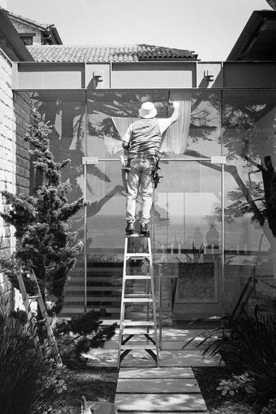 A black and white photograph of a person on a ladder running a squeegee across a broad glass surface, surrounded by more glass surfaces
