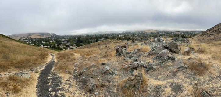 A panoramic view of a hilly landscape featuring dry grass, rocky terrain, and a pathway. In the background, a residential area is visible beneath a cloudy sky. The scene conveys a sense of natural beauty and tranquility.