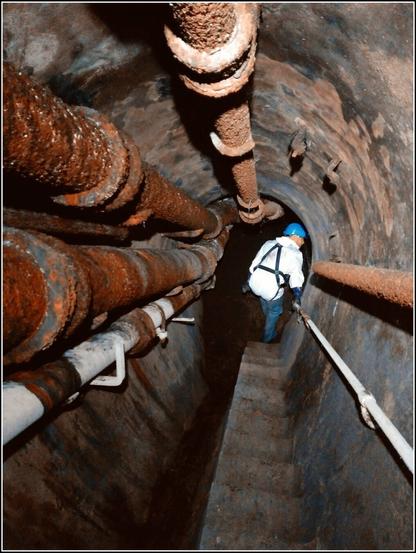 Un égoutier descendant un escalier en égout. Des canalisations sont fixées en voûte et sur les piédroits.