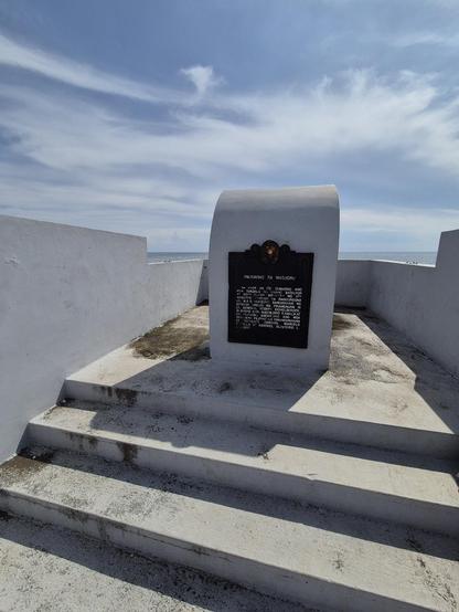 Historical marker mounted vertically on a concrete pedestal with a few steps before it and surrounded by a low concrete wall.