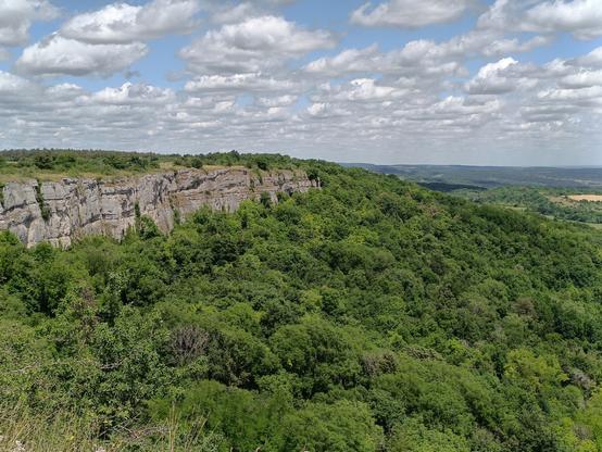 Photographie numérique couleur en format paysage.
A gauche, une falaise rocheuse avec des bosquets au-dessus et une forêt en contrebas. Le ciel est bleu et lumineux avec de nombreux nuages blancs.