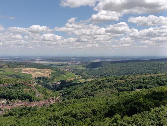 Même type de photo et de ciel, avec la suite du paysage à droite du précédent. Vue très large du bas du village, des forêts en contrebas de la falaise et de cultures.