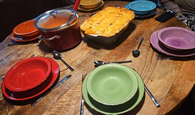 A round dinner table with six matching plates and bowls, each in a different color of the rainbow. In the middle is a pot of (bell pepper) soup and a big lasagne dish.