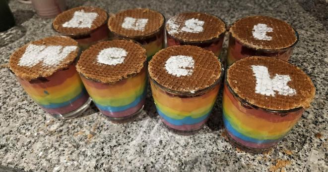 Eight glasses filled with six layers of yoghurt in the colors of the rainbow, topped off with a stroopwafel. On top, letters made out of powdered sugar spell out RustWeek.