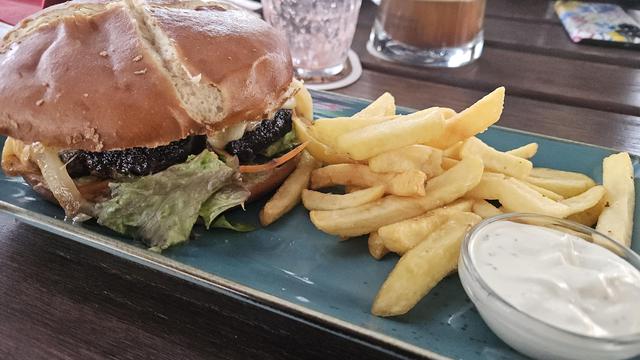 Photo of a burger and fries. The beef is fried very dark and hiding in a pretzel bun. Next to the fries is a tiny glass bowl is white sour creme. The plate is turquoise and rectangular. The table is made from dark wood. Glasses stand in the background.
