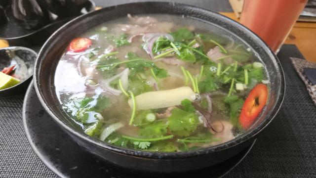 A big dark bowl full of a clear broth. Lots of cilantro swims in the broth. In between are bits of cooked beef visible. Two big pieces of chili are visible.
