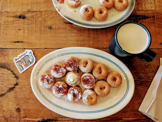 A wooden table with two plates of mini-doughnuts with different toppings, a mug of chai tea, and a sticker that says "You belong at Pips"