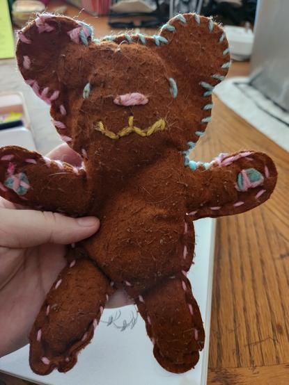a photo of a worn hand made teddy bear. he is brown and pink stitching goes all around the edges of the teddy bear. the pawpads are crudely attached and the eyes and nose are single loops.