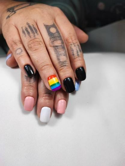 Manos con uñas recién pintadas en distintos colores. Una mano muestra uñas negras y una uña con la bandera LGBT en franjas horizontales. La otra tiene uñas en tonos rosado, blanco y celeste, evocando la bandera trans. Ambas manos tienen tatuajes visibles con taris. Fondo blanco liso