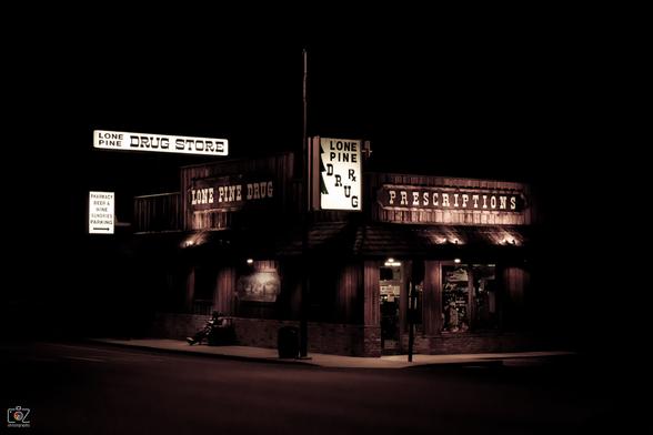 Dark photo in black and brown tones. It shows a single-storey wooden building in Western style at night, with a number of illuminated signs or inscriptions reading "Lone Pine Drug Store", "Pharmacy Beer & Wine Sundries Parking", "Lone Pine Drug" and "Prescriptions". A glass door with a sign "We have Ice Cream" and shop windows can be seen at the front. On the left side of the building, someone with a hat and outstretched legs is sitting on a bench.