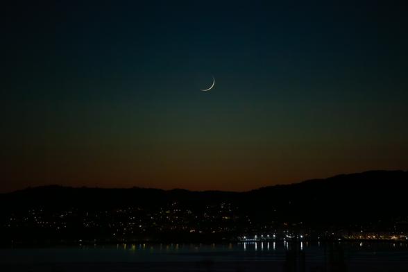 An almost new moon rises over the horizon in a dark picture, where you can still see the last traces of light after the sun set
