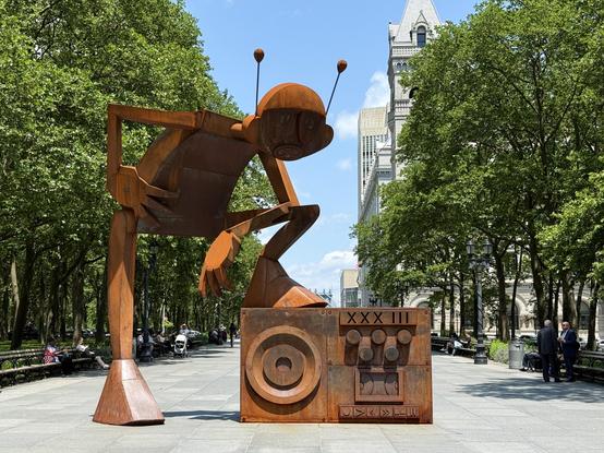 A rust colored statue of a robot with one leg up on a boom box. Downtown Brooklyn can be seen in the background with some trees and buildings.