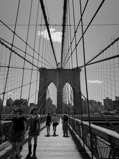 Black and white photo looking at the Brooklyn Bridge while walking across it. The path is made of wood, there are other people walking in front of us, and the metal fencing makes a spiderweb pattern around us.