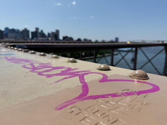 Hot pink graffiti writing on the side of the Brooklyn Bridge. Tall buildings of Brooklyn can be seen in the background agains a blue sky.