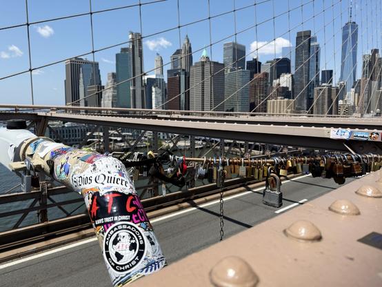 Some love locks on a support for a light that is covered in colorful stickers. Looking over the road portion on the bridge, you can see tall building of New York City in the background with blue sky and a few small white clouds.