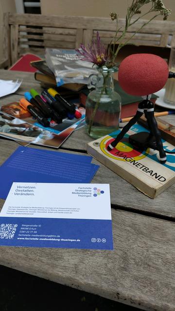 Ein Flyer der Fachstelle Strategische Medienbildung Thüringen, ein Mikrofon und ein Magnetband liegen auf dem Tisch