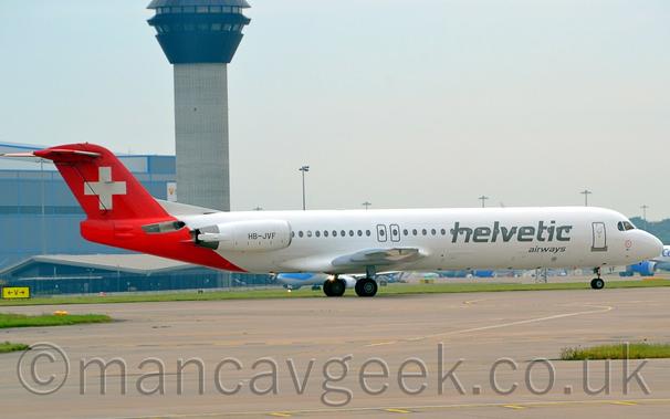 Side view of a twin engined jet airliner taxiing from left to right.
The plane is mostly white, with large grey "Helvetic" titles covering the forward passenger cabin windows, above smaller grey "Airways" text.
The registration "HB-JVF" is on the white engine pods, mounted on the sides of the rear fuselage.
The rear fuselage and tail are red, with a white cross in the middle.