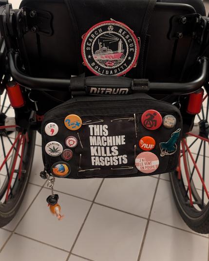 Photos de l'arrière de mon fauteuil roulant, en vue rapproché. Sur le dossier un patch avec un bateau et le mot search rescue solidarity.
Dessous, une pochette avec un patch This Machine Kills Fascists et une dizaine de badge (Ubuntu, cannabis, geek et libriste).