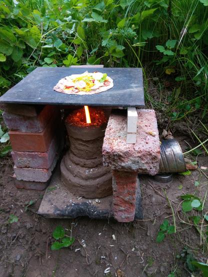Ein kleiner Lehmigen steht vor grünem Hintergrund. Herumgebaute Ziegel halten eine Stahlplatte über die Flamme. Auf der Platte liegt ein belegtes Fladenbrot.
