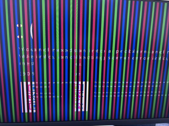 Photo of a computer monitor displaying a Windows Blue Screen of Death; something has gone wrong with either the OC or the display, though, as the screen is divided into vertical stripes of repeating red, blue, and green, with thin regions of black between them. The frowning face emoticon of the BSOD is visible. The same bug causing the vertical color stripes has also garbled the message text, but unscrambled it would say, “Your PC ran j to a problem and needs to restart.” A distorted QR code is also visible.