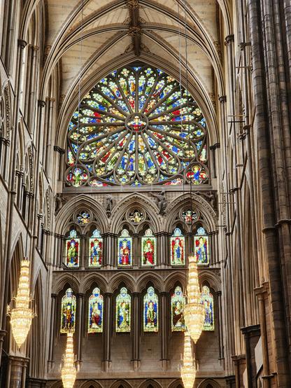 Large Rose window with predominantly yellow, red and blue glass below a ribbed-vaulting ceiling. Below the Rose window are two rows of smaller windows, each depicting a saint or biblical figure.