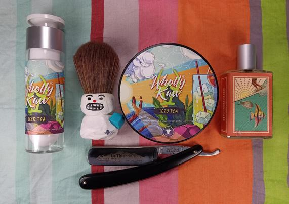 (Le Chat Mistral description) 

The image displays a collection of grooming items arranged on a striped, multicoloured background. On the left, there is a bottle of Wholly Kaw Iced Tea-scented shaving cream. Next to it, a small, cute, white figure with a brush-like top stands out. In the centre, there is a round tin of Wholly Kaw Iced Tea-scented balm. To the right of the tin, there is a bottle of perfume with a label featuring tropical fish. At the bottom, there is a black straight razor with "Le Thiernois" engraved on the blade. The items are colourful and neatly arranged.