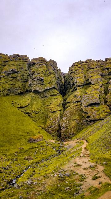 A narrow dirt trail winds through a lush green valley toward a dramatic rock formation split by a deep crevice. The steep cliffs are covered in moss and vegetation, with jagged layers of stone creating a striking texture. A small stream flows alongside the path, originating from the mouth of the rocky gap. A few tiny hikers near the crevice provide a sense of scale, emphasizing the towering cliffs. The sky is overcast, casting a soft light over the vivid green landscape.