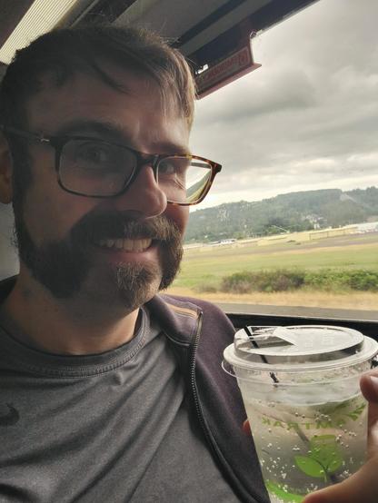 A selfie with a Gin and Tonic in my train seat. The drink is in a plastic cup with a green Amtrak logo, a lid, a thin straw and a sunken lime wedge. Outside the windows it is overcast. There's light industrial buildings and tree covered hills. It all looks a bit blurry with the movement of the train.