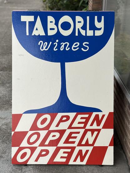 A handpainted A-frame reads “TABORLY WINES OPEN OPEN OPEN”. The sign features an abstract illustration of a deep blue goblet or stemmed glass on a red and white checked surface that resembles a tablecloth.