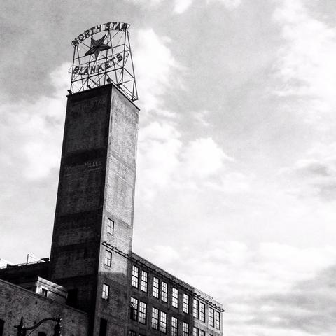 A tall tower in an old factory rises into the sky with an old sign on top that reads, North Star Blankets. A large star is centered in the sign with words around it. A black and white photograph