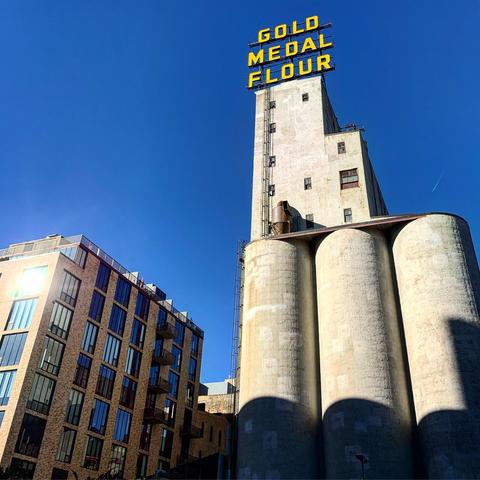A tall grain elevator with three white cylindrical towers stands before a blue sky. Atop is a large yellow sign that has the words Gold Medal Flour.