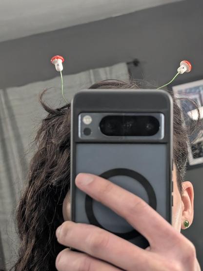 A picture of this enby taking a zoomed in selfie. Their face is obscured save for those high cheekbones. They're wearing grassy green plugs in their ears. Messy curls falling in waves down one side. The mushroom sprouts atop her head have moved. They have spotted red tops and on the white stems they have grown a googly eye each. And the eyes are on you.