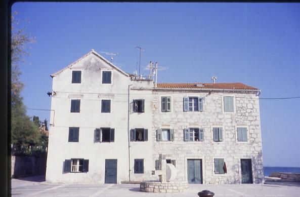 Large whitewashed stone building against a blue sky and sea, morning or midday. One section has four stories, two windows each, except the ground floor has one window and a door. The other section is three stories tall and has 12 windows and two doors. Most of the windows are covered by double shutters, dark blue and light blue. It has an orange clay tile roof. There are narrow cylindrical chimneys/vents and television antennae on the roof.
The building is on a vacant plaza of white stone and in the center is a construction that I am guessing is a water fountain or well. A low circular wall of (yes) white stone, and from the center is a large vertical beam with a horizontal protrusion. Behind this is a large disk on edge, which I am guessing is some kind of water wheel for pumping water. I could be very wrong.