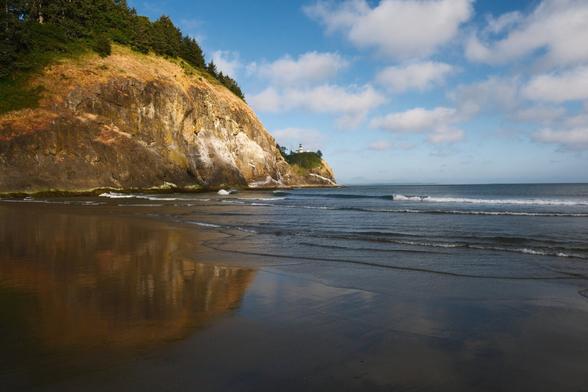 On the top left, a large hill with a small hill coming off of it, with a small lighthouse on top next to a partially cloudy sky on the top right. The bottom half of the image is small waves gently rolling from the right onto a beach on the left. The wet beach is reflecting the hill from the top of the image.