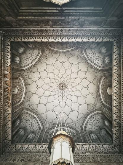 View up into the ceiling of the mosque. An incredibly detailed mandala pattern seems to pull one up into higher realms, while a pendant light is suspended right from its center, bringing light into the earthly halls of this ancient place of worship.