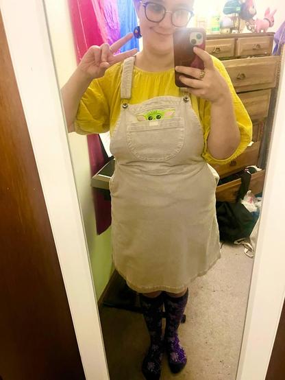 mirror selfie of a young white person with short brown hair and glasses. i'm wearing a corduroy overall dress with embroidery in the pocket meant to look like grogu/baby yoda/the child. underneath the dress is a yellow shirt with puffed sleeves. my earrings are paper quilling rainbow flowers and my knee socks have the griffon-and-chalice motif of the grey wardens from dragon age.