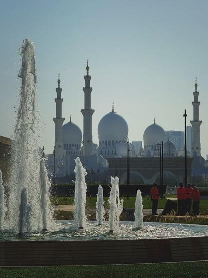 A group of workers in red shirts, wedged in between an opulent water fountain in the foreground and the white domes of a large mosque in the background