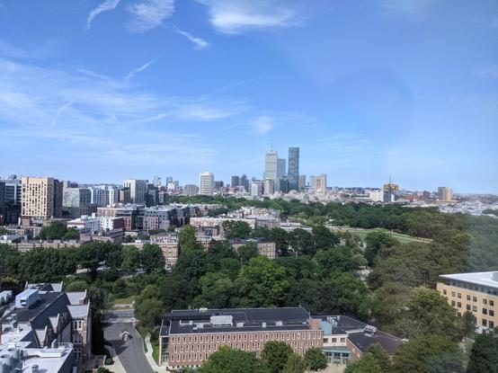 View from high floor into Boston with skyscrapers in the distance, green trees below and blue sky with a few wispy clouds