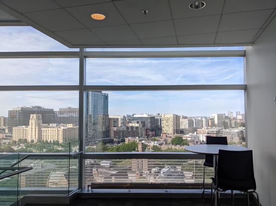 View from 11th floor lobby through windows on wall of building onto buildings in the distance. Small round table with 2 chairs to the right