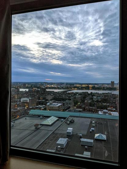 View from high floor window onto buildings low down and Charles river in distance and cloud covered sky at dusk