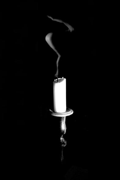 A vertical, black and white image of an unlit candle standing in the lower half of the image. At the upper half stands a naked woman and only some parts of her body are visible, seen in profile. She has bent knees, leaning forward and holds one arm up behind her. The only visible parts of her body is her bottom, the back of her legs and her arm, the rest of her body lays in shadow. Since she is standing in the background she is blurry which makes her body look like the smoke curling upwards after the extinguished flame.