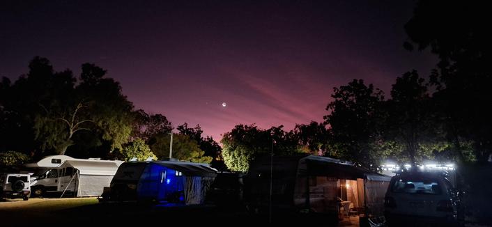 Red pink glowing sky when partial moon and one star.  Caravans and other camping sights glowing with their lights. Shadows of trees against the sky.