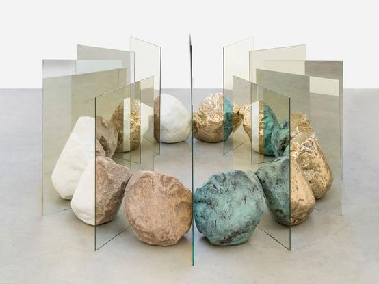A collection of large granite rocks form a connected circle with tall mirrors between them. They are all touching to form a solid ring. The mirrors appear to be glass, revealing rocks that are split down the middle, half one color and half another. They distort reality in being unable to define the sheets of glass as transparent or reflection.