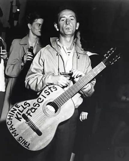 Iconic black and white photo of folk musician Woody Guthrie looking at the camera, holding his acoustic guitar that has the words, THIS MACHINE KILLS FASCISTS written on it.