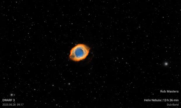 A bright blue disk of nebula with just a few stars, surrounded by a yellow ring and then an orange outer ring with two darkened 'ears'. In the bottom corners are the captions "Rob Masters" "Dwarf3" "2025.06.28 09:17" "Helix Nebula | 13h 36 min" and "Duo Band".