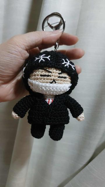 4-inch-tall crochet doll keyring of Suga from the Hope on the Stage Final concert. Suga doll is wearing a black beanie with snowflake designs on the brim, a white mask, and a black cardigan and pants.