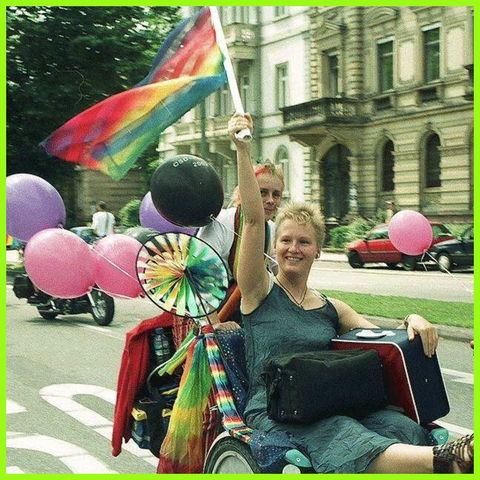 Farbfoto: 2 Frauen auf einer Art Fahrradrikscha, die Frau vorn trägt eine Regenbogenfahne, das Fahrrad ist geschmückt mit Ballons und anderen Utensilien